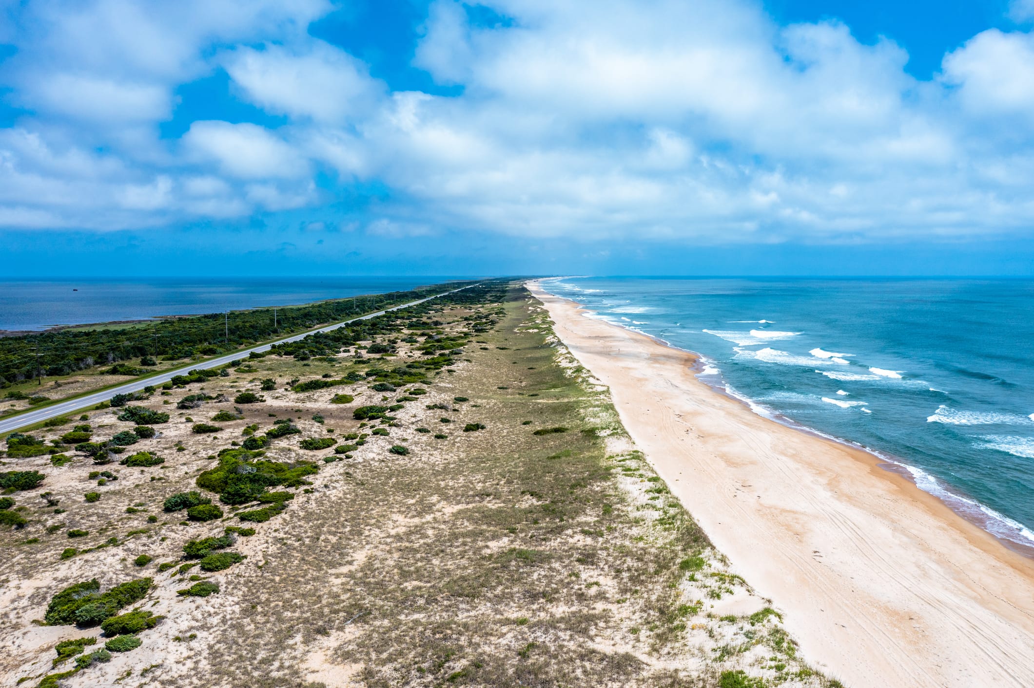Aerial view of a long, sandy beach with gentle waves and a road parallel to the coastline under a partly cloudy blue sky.