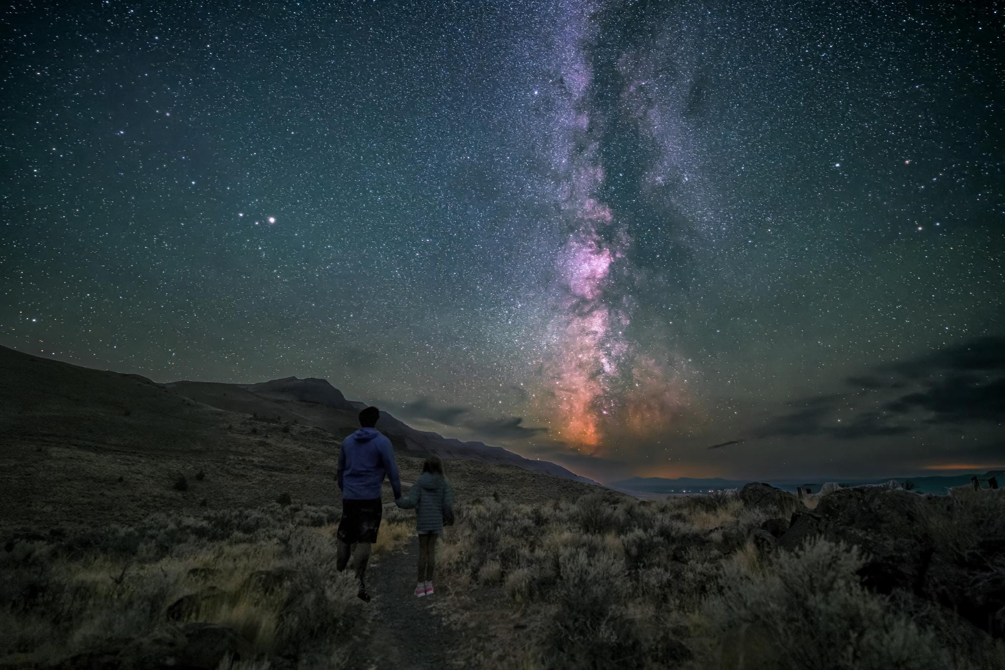 A couple walks hand in hand on a path under a vibrant, star-filled night sky in the Oregon Outback.