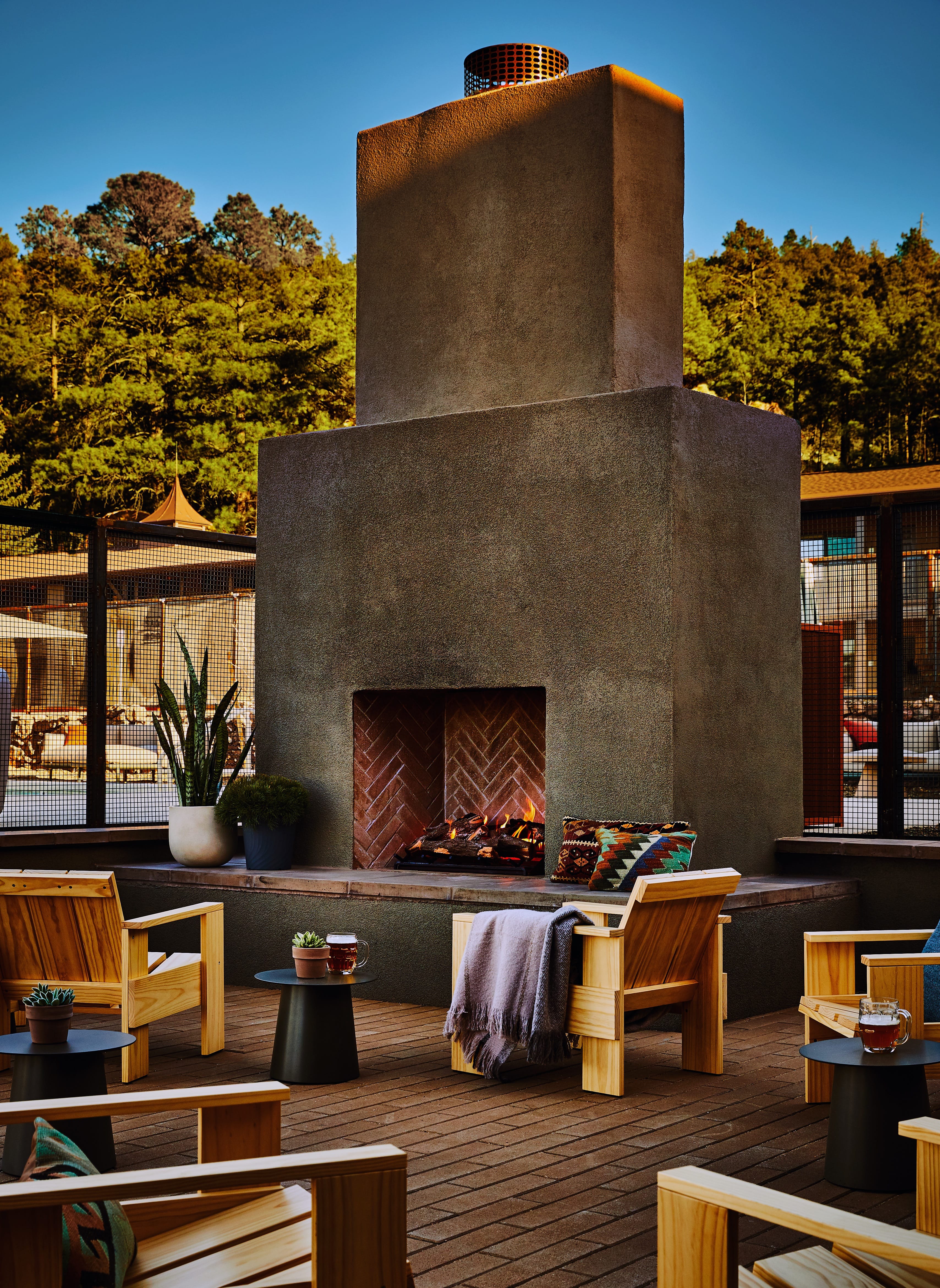Outdoor patio with a large stone fireplace, wooden chairs, small tables, and potted plants, surrounded by a fence and trees under a clear sky.