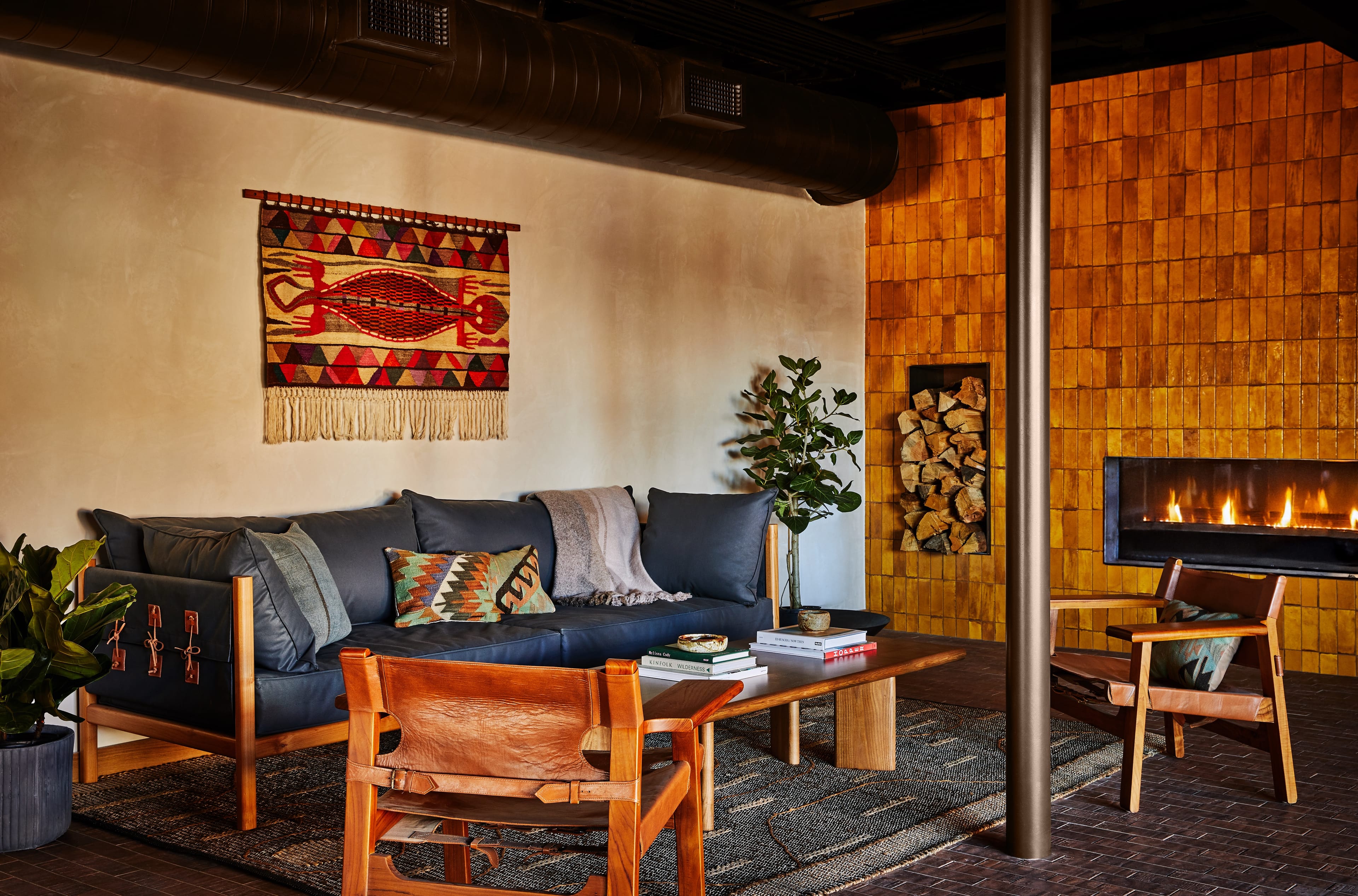 Cozy living room with a dark sofa, wooden chairs, a wall tapestry, a modern fireplace, and a stack of firewood. Warm lighting enhances the ambiance.