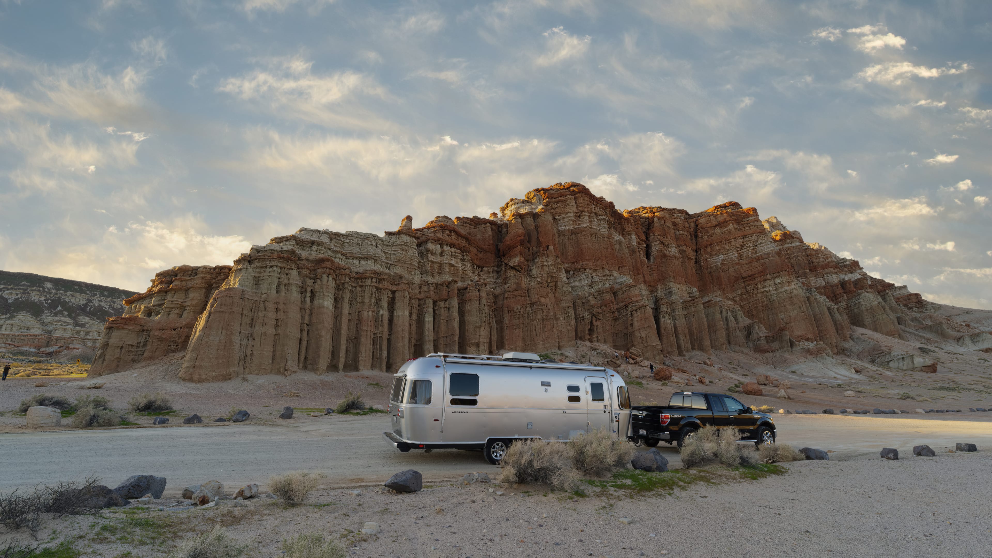 A silver Airstream trailer and a black vehicle are parked near dramatic red rock cliffs under a cloudy sky in a desert landscape.